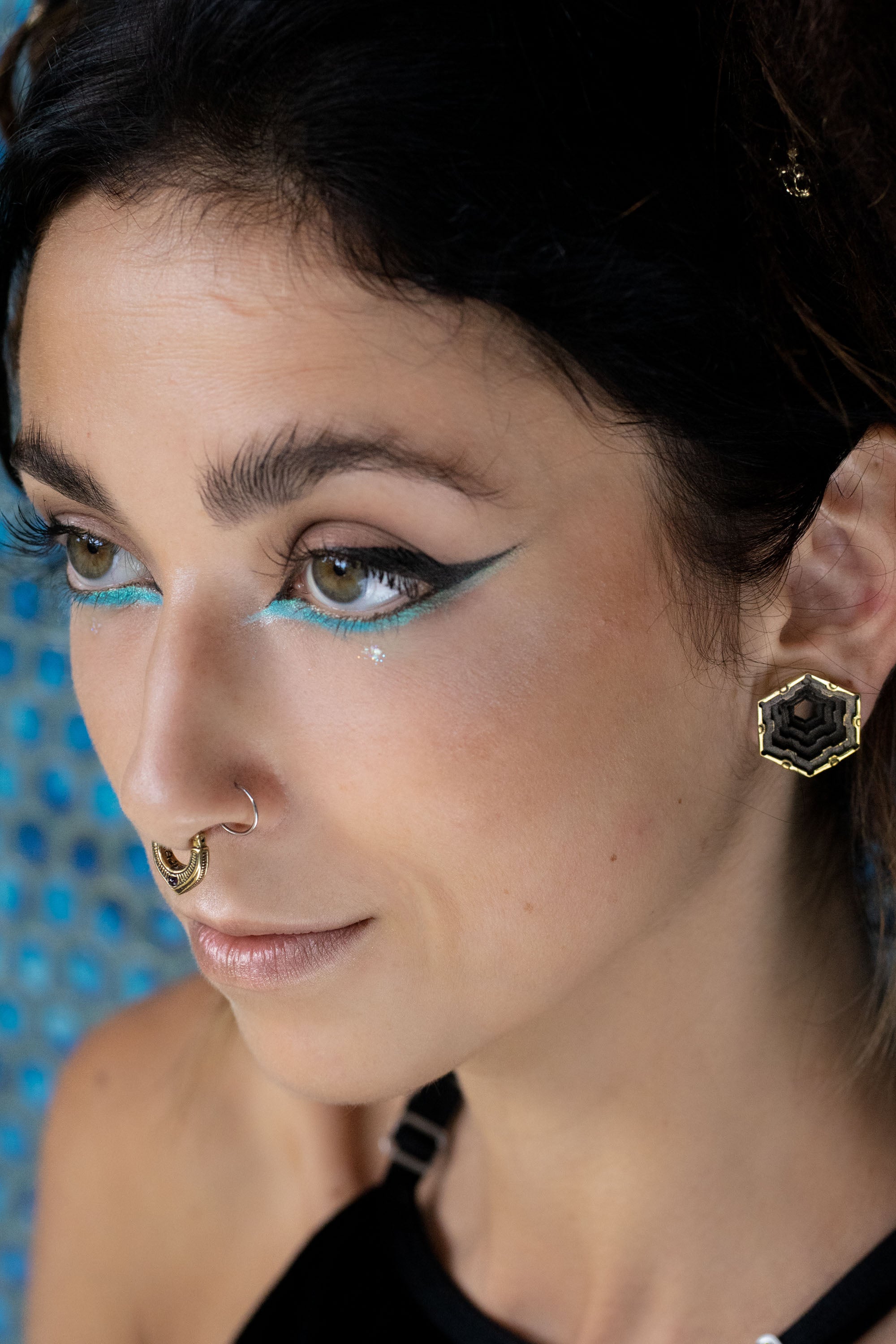 Close-up of a woman with bold turquoise and black winged eyeliner, wearing a gold septum ring and hexagonal black-and-gold earrings. Her expression is serene, and her brown eyes are softly gazing to the side.